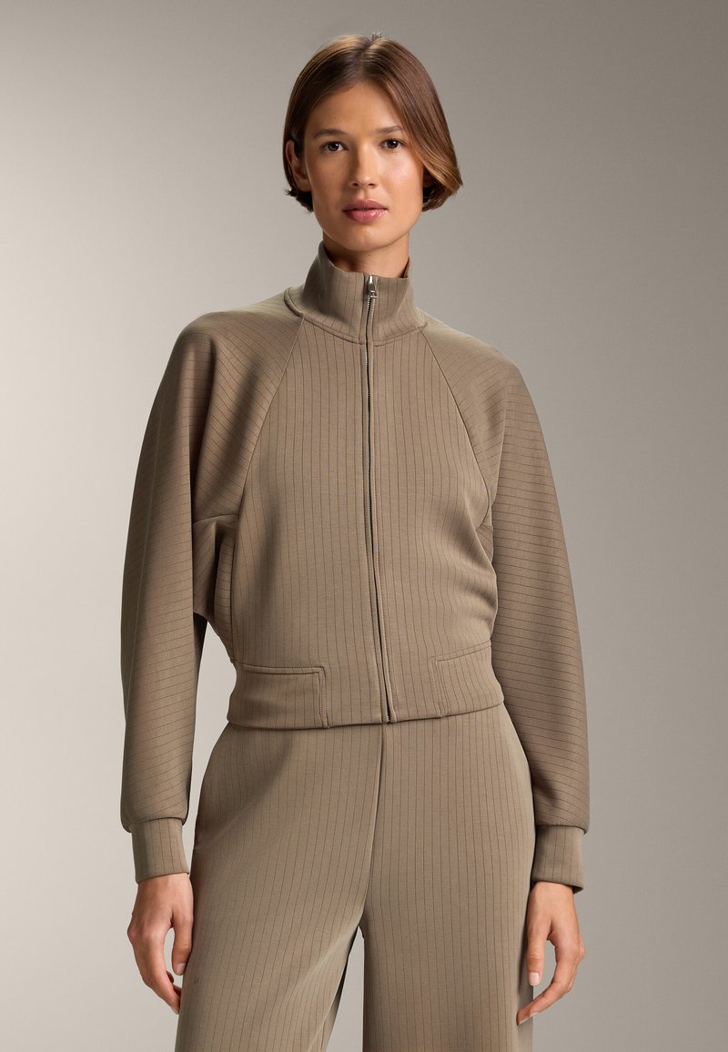 Hnedá rebrovaná bunda na zips s vysokým golierom, ktorá má raglánové rukávy a predné vrecká. Kombinovaná s vhodnými rebrovanými nohavicami.