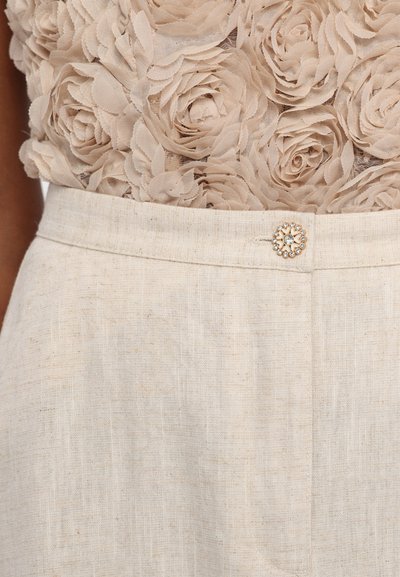 Beige topp med blomstermønster og 3D-stoffroser, kombinert med lyse linbukser som har en dekorativ juvelknapp i livet.