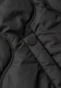 Czarna ocieplana kamizelka z gładką fakturą, pikowanym wzorem i okrągłą czarną metką z wytłoczonym napisem.