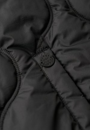 Gilet rembourré noir avec une texture lisse, un design matelassé et une étiquette circulaire noire avec un lettrage en relief.