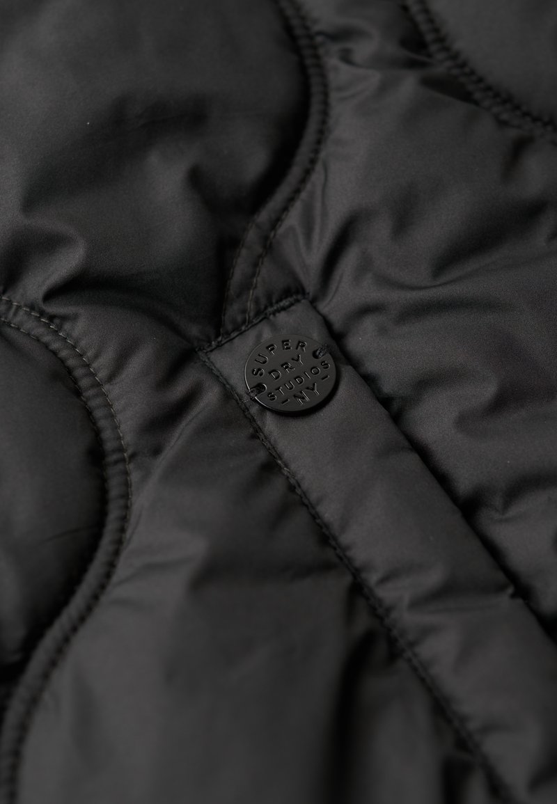 Schwarze gepolsterte Weste mit glatter Oberfläche, gestepptem Design und einem runden schwarzen Logoanhänger mit geprägtem Schriftzug.