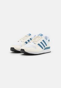 Pár bílých a modrých tenisek Adidas s detaily z prodyšné síťoviny a semiše, zavázané a postavené vedle sebe na bílém pozadí.