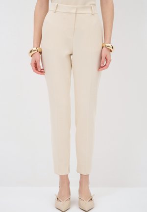Beige tailored trousers with a slim fit, featuring a smooth texture, front pleats, and belt loops. Paired with beige pointed-toe heels.
