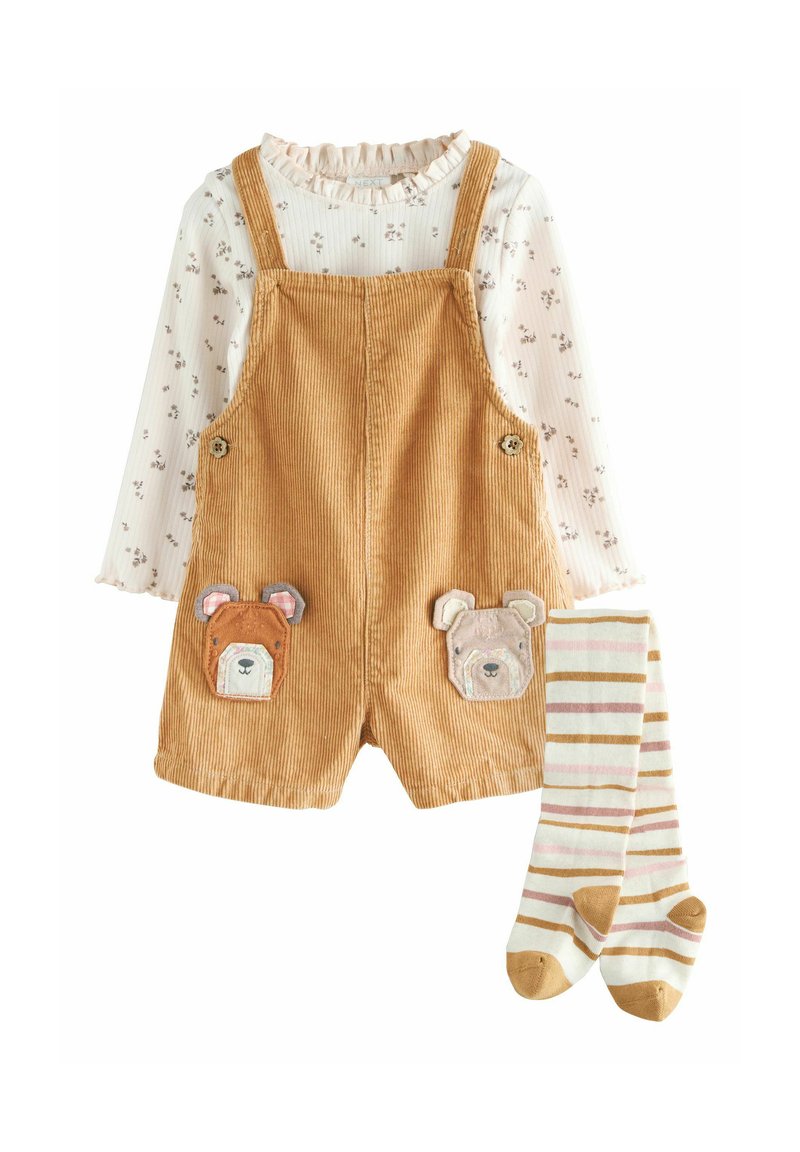 Pantaloni salopette in velluto a coste marrone con tasche applique a forma di orso, abbinati a una maglietta bianca a maniche lunghe con stampa floreale e calze a righe alte fino al ginocchio.