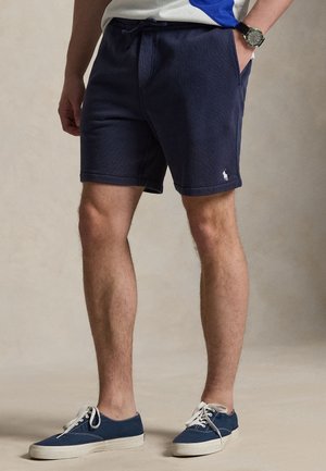 Man draagt marineblauwe korte broek met logo op linkerbeen, horloge aan linkerpols en blauwe canvas schoenen met witte veters op een neutrale achtergrond.