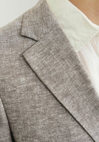 Blazer de lino gris con tejido texturizado, que presenta un cuello afilado y costuras visibles. La camisa debajo es de color claro y tiene un acabado suave.