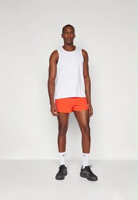Camiseta deportiva blanca sin mangas, pantalones cortos naranjas, zapatillas negras y calcetines blancos. Diseño simple con un corte ceñido y una marca mínima.