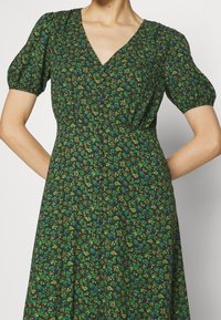 Robe fleurie vert foncé avec manches courtes bouffantes, décolleté en V et design à boutons sur le devant. Présente de petites fleurs jaunes et vertes.