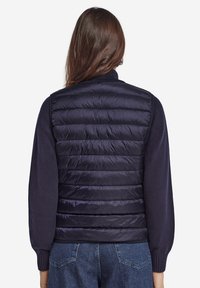 Marinblå puffer-väst med quiltad textur, hög krage och figursydd nederkant, kombinerad med en marinblå långärmad topp och denimbyxor.