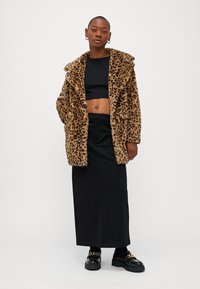 Kapa od umjetnog krzna s uzorkom leopard, širokim ovratnikom, dvjema bočnim džepovima i ležernim krojem, nosi se preko crnog topa i crne suknje.