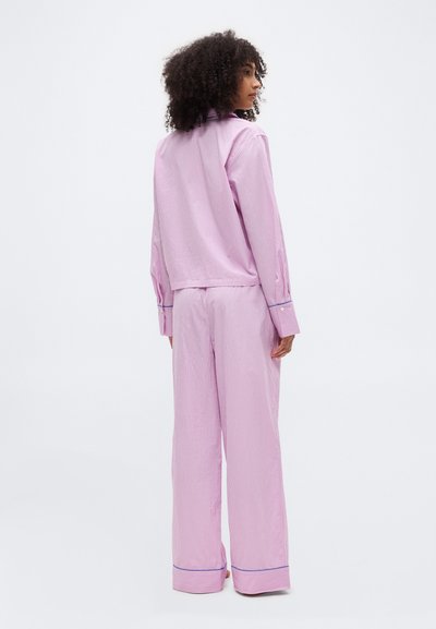 Femme aux cheveux bouclés vue de dos, portant une chemise à manches longues rayée rose et blanche et un pantalon à jambes larges avec des bordures bleues aux poignets et aux ourlets.
