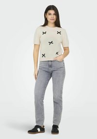 Crème korte mouwen sweater met zwarte X-patroon accenten, gecombineerd met grijze denim jeans en zwarte sneakers met witte strepen.