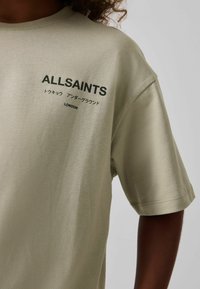 Hele roheline lühikeste varrukatega T-särk, mis on valmistatud pehmest kangast, musta tähestiku "ALLSAINTS" ja mitmekeelse tekstiga, lõdva konstruktsiooniga disainiga.