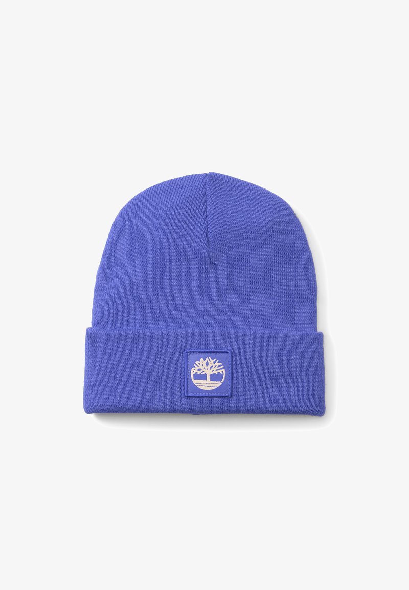 Lila stickad mössa med uppvikt kant, prydd med en fyrkantig lapp med en vit trädlogo. Mjuk textur, enkel design.