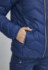 Modrá puffová bunda s chevronovým prešívaním, vybavená strieborným zipsom a bočnou kapsou. Mäkký, hladký materiál a rebrované manžety.