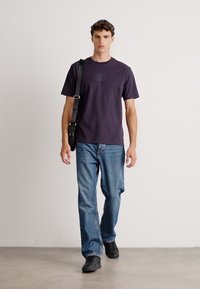 Fiatal férfi sétál a beltérben, fekete pólót, kék farmernadrágot, fekete cipőt és fekete válltáskát viselve.