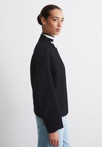 Sort bomberjakke med ribbet krave, lange ærmer og sidelommer, iført over en sort-hvid stribet turtleneck. Lyseblå jeans er synlige.