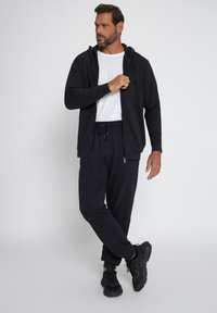 Sudadera negra con cremallera y capucha ajustable, combinada con pantalones de chándal negros. El conjunto incluye una camiseta blanca debajo y zapatillas negras.