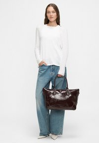 Brązowa skórzana torebka o sztywnym kształcie, z podwójnymi uchwytami i srebrnymi akcentami. Modelka nosi białą bluzkę z długim rękawem i szerokie dżinsy.