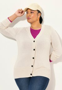 Beige stickad cardigan med svarta knappar, v-ringad design, bärs över en lila skjorta, ihop med en beige keps. Har ribbade ärmslut och nederkant.