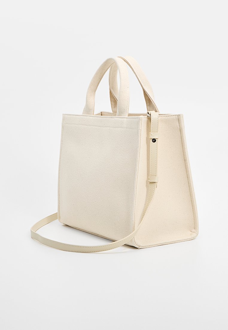 Sacs fourre-tout en toile beige avec des doubles poignées courtes et une bandoulière amovible, présentant un design structuré et des détails de couture minimaux.