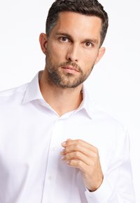 Camicia bianca con colletto a punta, caratterizzata da una superficie liscia e un leggero lucido; vista su una persona che aggiusta il colletto con una mano.