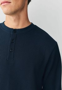 Mężczyzna w ciemnoniebieskiej koszulce henley z dzianiny w wafle, z trzema guzikami, widoczny od szyi do ramienia na jednolitym jasnym tle.