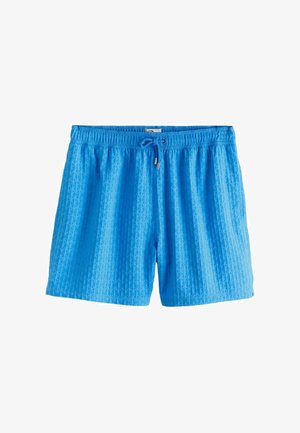 Bărbat fără tricou cu păr creț poartă pantaloni scurți de baie albaștri, sandale negre și un colier, stând în fața unui perete crem texturat.