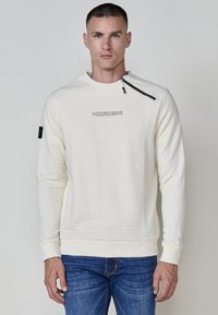 Sudadera de punto color crema claro con superficie texturizada, cuello redondo, mangas largas y un detalle con cremallera en el hombro izquierdo; logo impreso en el pecho.