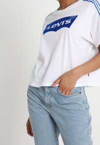 Camiseta de algodón blanca con logo azul de Levi's, mangas cortas y rayas azules en los hombros. Combinada con jeans azul claro.