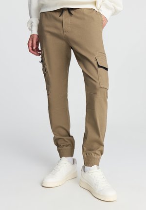 Pantaloni cargo khaki con polsini elasticizzati, dotati di tasche laterali con zip e vita regolabile con cordino, abbinati a sneakers bianche.
