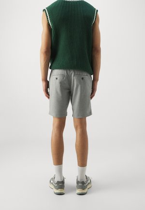 Pull sans manches en maille verte avec des accents blancs, associé à un short gris clair. Chaussures de sport grises avec des accents clairs et chaussettes blanches.