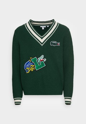 Pull en maille vert foncé à col V, col et poignets rayés blancs, avec deux patchs crocodiles colorés sur le devant.