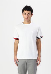Hvid kortærmet t-shirt med rund hals, prydet med marineblå og røde striber på ærmerne og et lille logo på brystet.