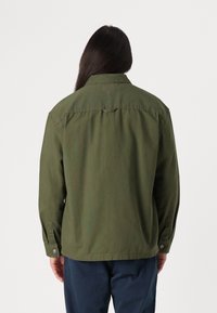 Veste vert olive, en coton, coupe ample, manches longues, poignets à boutons, col pointu, détail de yoke au dos, design simple.