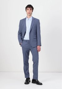 Helles blaues Anzug aus strukturiertem Stoff, bestehend aus einem einreihigen Jacket, passenden Hosen und einem hellblauen Hemd darunter. Schwarze Schuhe.