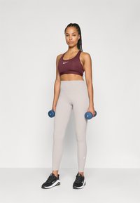 Atletisch outfit met een wijnrood sportbeha, lichtgrijze high-waisted leggings en zwarte sneakers, terwijl ze blauwe dumbbells vasthoudt.