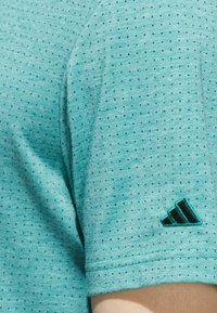 Turkoosi lyhythihainen paita, jossa on pieniä mustia pisteitä ja teksturoitu kangas. Paidassa on brodeerattu musta Adidas-logo hihassa.