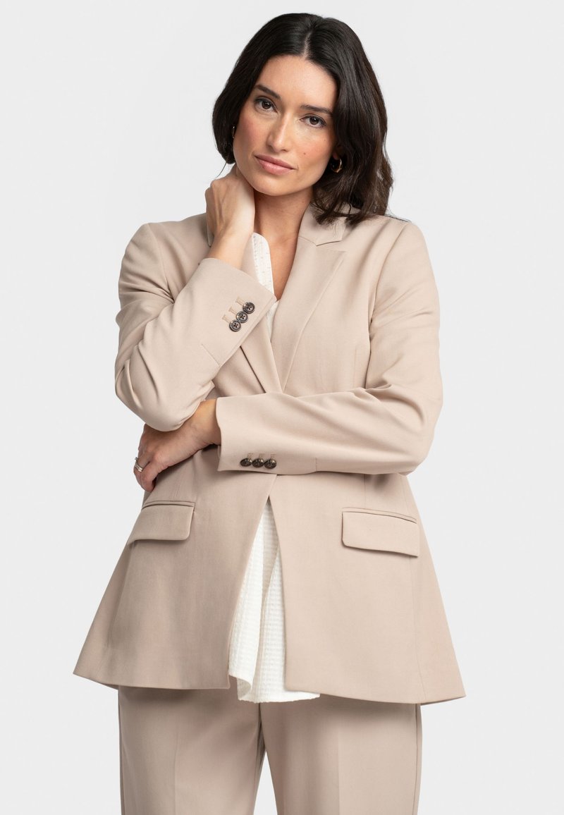 Woman with dark hair wearing a beige suit jacket and pants, posing with one hand on her neck and the other crossed over her waist.