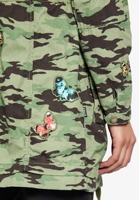 Kamouflagejacka i grönt och svart, prydd med broderade fjärilspatchar i blått, orange och rosa, med knappar som accenter.