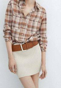 Donna che indossa una camicia a quadri marrone con bottoni, infilata in una mini gonna in maglia a coste beige, con una ampia cintura marrone.