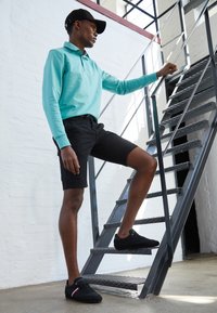 Młody mężczyzna w czapce, turkusowej, długiej koszuli, czarnych szortach i czarnych trampkach wspinający się po metalowych schodach wewnątrz budynku.