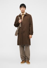 Καφέ μάλλινο παλτό με γιακά, εμπρόσθιες τσέπες και κούμπωμα με κουμπιά, φορεμένο πάνω από μπεζ πουλόβερ και ανοιχτό καφέ παντελόνια, με γκρι τσάντα χιαστή.