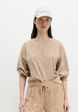 Kobieta w beżowej bluzie z frotty i dopasowanych szortach z troczkiem oraz białej czapce Marc O’Polo, stojąca na białym tle.