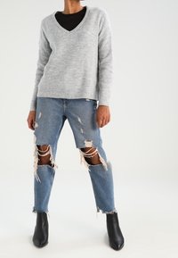 Jasnoszary sweter z dekoltem w serek, luźny krój; niebieskie dżinsy z dużymi przetarciami na kolanach; czarne botki za kostkę; gładka faktura i casualowy design.