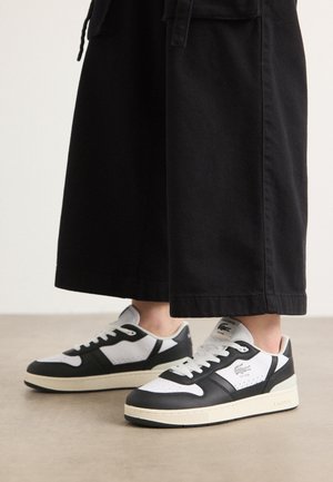Personne portant des baskets basses Lacoste noires et blanches avec un pantalon noir à jambes larges coupé au-dessus de la cheville.