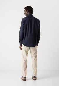 Camisa de manga larga azul marino de tela de algodón, con puños abotonados y un dobladillo curvado, combinada con pantalones color crema y zapatos marrones.