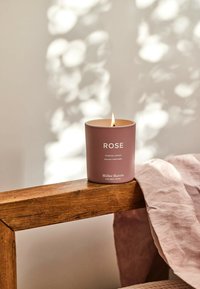 Rosenduftkerze in einem matten mauvefarbenen Behälter mit weißem Text; auf einer Holzoberfläche mit einem hellrosanen Tuch, das in der Nähe liegt.