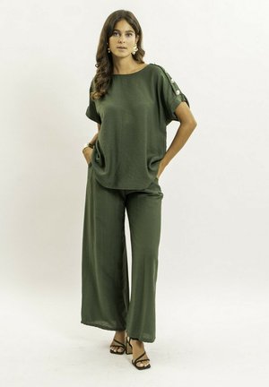 Femme debout, mains dans les poches, vêtue d'un haut fluide à manches courtes olive avec boutons et d'un pantalon large assorti, talons noirs.