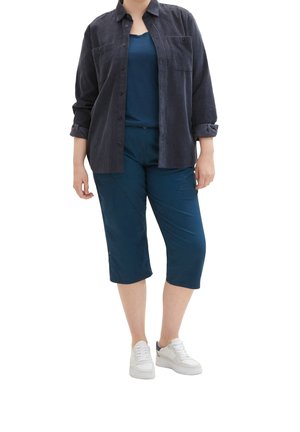 Marineblauwe ribstof blouse met knopen over een donkerblauwe tanktop, gecombineerd met teal crop pants en witte sneakers. Kenmerken zijn onder andere zakken en manchetbelangrijk.